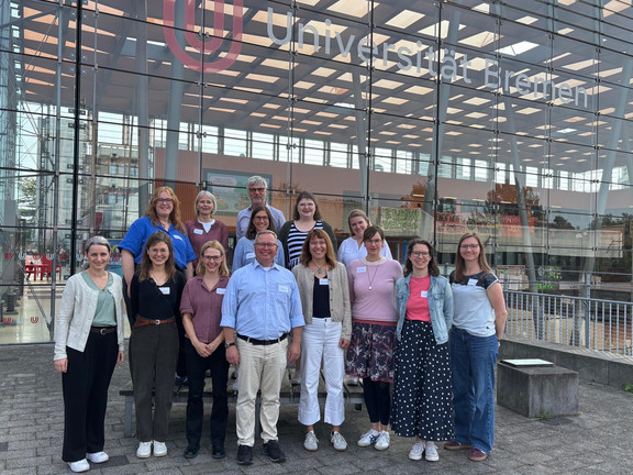 Gruppenfoto von 13 Personen vor dem verglasten Eingangsbereich der Universität Bremen. Die Gruppe steht in zwei Reihen dicht beieinander und blickt lächelnd in die Kamera. Im Hintergrund sind durch die große Glasfassade Teile des Universitätsgebäudes zu sehen. Oberhalb der Gruppe befinden sich das Logo und der große Schriftzug „Universität Bremen“.