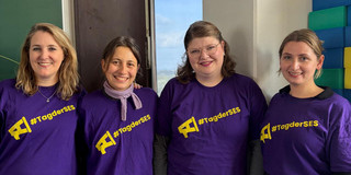Vier lächelnde Personen stehen nebeneinander in einem hellen Raum und blicken in die Kamera. Alle tragen lilafarbene T-Shirts mit einem gelben Megafon-Symbol und dem Schriftzug „#TagderSES“. Im Hintergrund sind eine grüne Tafel, ein hohes Fenster und gestapelte bunte Schaumstoffbausteine zu sehen. Die Gruppe steht eng beieinander und posiert für ein gemeinsames Foto.