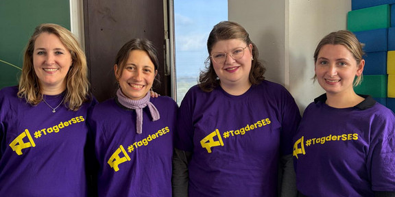 Vier lächelnde Personen stehen nebeneinander in einem hellen Raum und blicken in die Kamera. Alle tragen lilafarbene T-Shirts mit einem gelben Megafon-Symbol und dem Schriftzug „#TagderSES“. Im Hintergrund sind eine grüne Tafel, ein hohes Fenster und gestapelte bunte Schaumstoffbausteine zu sehen. Die Gruppe steht eng beieinander und posiert für ein gemeinsames Foto.