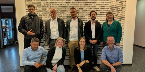 Gruppenfoto von neun Personen bei einem Abschlusstreffen an der TU Dortmund. Fünf Personen stehen nebeneinander vor einer Wand mit hellen, rechteckigen Mustern, vier Personen knien oder hocken davor. Alle blicken zur Kamera und lächeln. Die Gruppe steht in einem Innenbereich mit dunklem Boden und hellen Wänden.