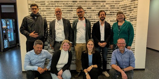 Gruppenfoto von neun Personen bei einem Abschlusstreffen an der TU Dortmund. Fünf Personen stehen nebeneinander vor einer Wand mit hellen, rechteckigen Mustern, vier Personen knien oder hocken davor. Alle blicken zur Kamera und lächeln. Die Gruppe steht in einem Innenbereich mit dunklem Boden und hellen Wänden.