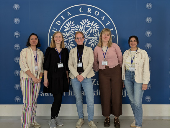 Five people are standing in front of a logo whose lettering is not fully recognizable. They are wearing name badges and looking into the camera.