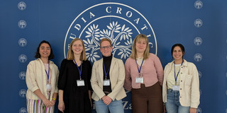 Five people are standing in front of a logo whose lettering is not fully recognizable. They are wearing name badges and looking into the camera.