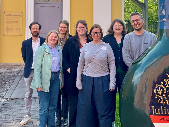 Sieben Menschen stehen nebeneinander vor einem Gebäude. Sie gucken alle in die Kamera.