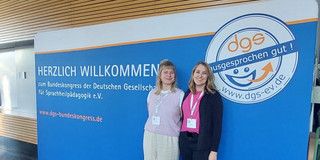 Das Foto zeigt Dr. Eva Wimmer und Pauline Schröder, die nebeneinander vor einer großen blauen Veranstaltungswand stehen und in die Kamera lächeln. Beide tragen Namensschilder an Schlüsselbändern. Auf der Wand steht „Herzlich Willkommen zum Bundeskongress der Deutschen Gesellschaft für Sprachheilpädagogik e.V.“. Rechts ist das große Logo der Gesellschaft mit dem Schriftzug „dgs – ausgesprochen gut!“ zu sehen. Die Aufnahme vermittelt den Kontext eines wissenschaftlichen Fachkongresses.