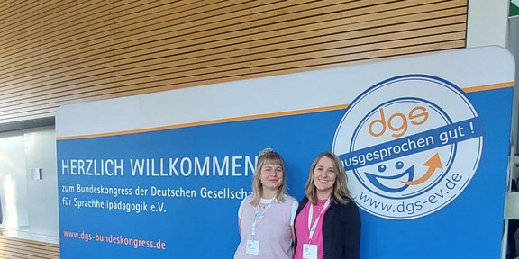Das Foto zeigt Dr. Eva Wimmer und Pauline Schröder, die nebeneinander vor einer großen blauen Veranstaltungswand stehen und in die Kamera lächeln. Beide tragen Namensschilder an Schlüsselbändern. Auf der Wand steht „Herzlich Willkommen zum Bundeskongress der Deutschen Gesellschaft für Sprachheilpädagogik e.V.“. Rechts ist das große Logo der Gesellschaft mit dem Schriftzug „dgs – ausgesprochen gut!“ zu sehen. Die Aufnahme vermittelt den Kontext eines wissenschaftlichen Fachkongresses.