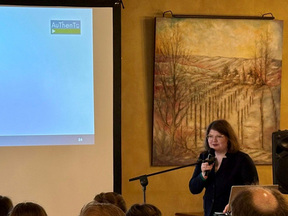 Eine Frau steht vor einer Präsentation mit einem Mikrofon in der Hand. Sie spricht zu Personen, die vor ihr sitzen.